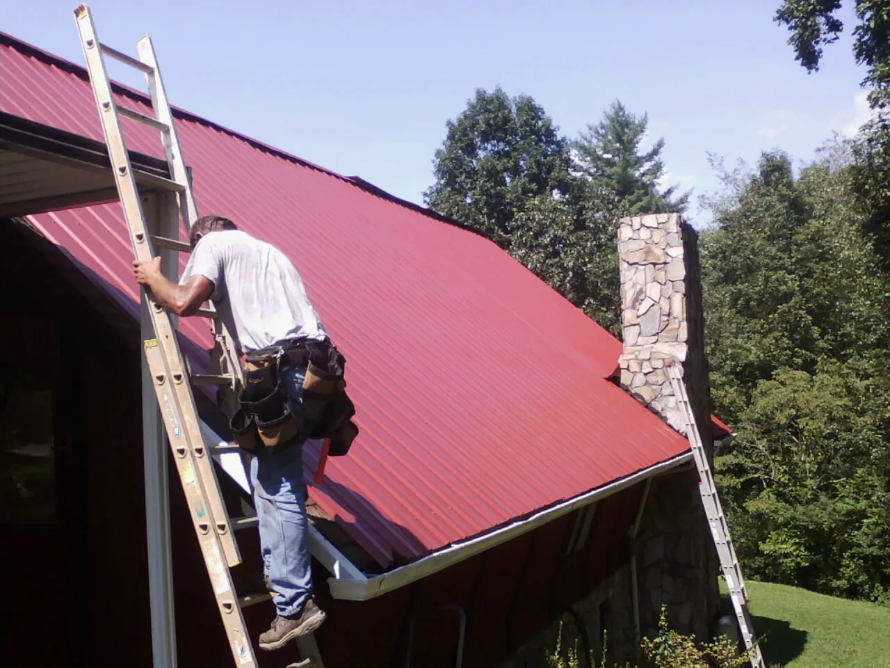 Roofer on a ladder beside a red metal roof and stone chimney — Soffit & Fascia Repair by Roofing Brothers in Bath Springs